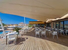 Zona pranzo all'aperto illuminata dal sole, con tavoli e sedie, una piscina e vista sugli edifici sullo sfondo.