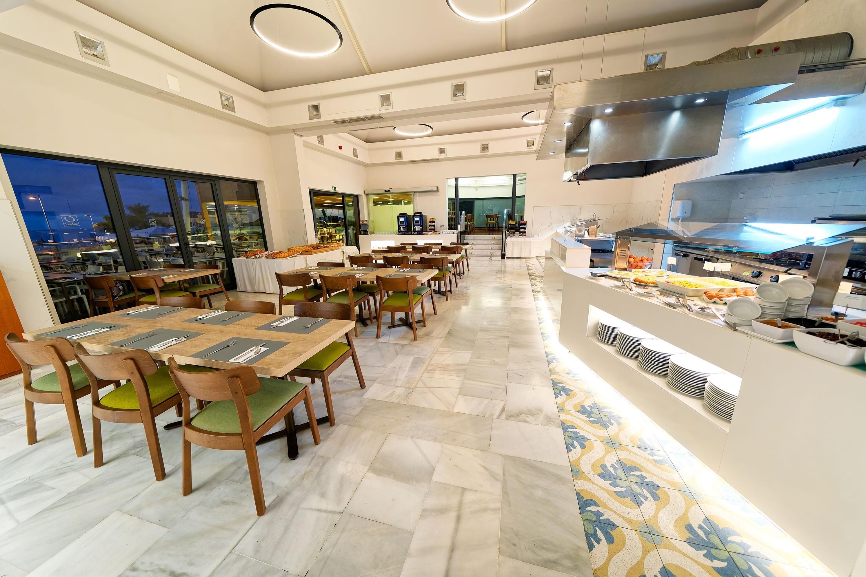Un comedor amplio con varias mesas y sillas, un mostrador de buffet y vistas a la ciudad a través de grandes ventanales.