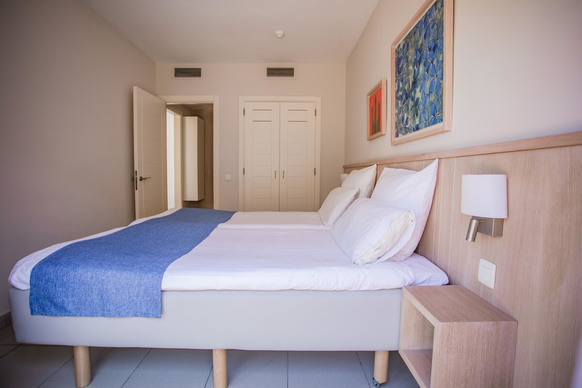 A neatly made double bed with a blue throw blanket, a bedside table with a lamp, and a painting on the wall.