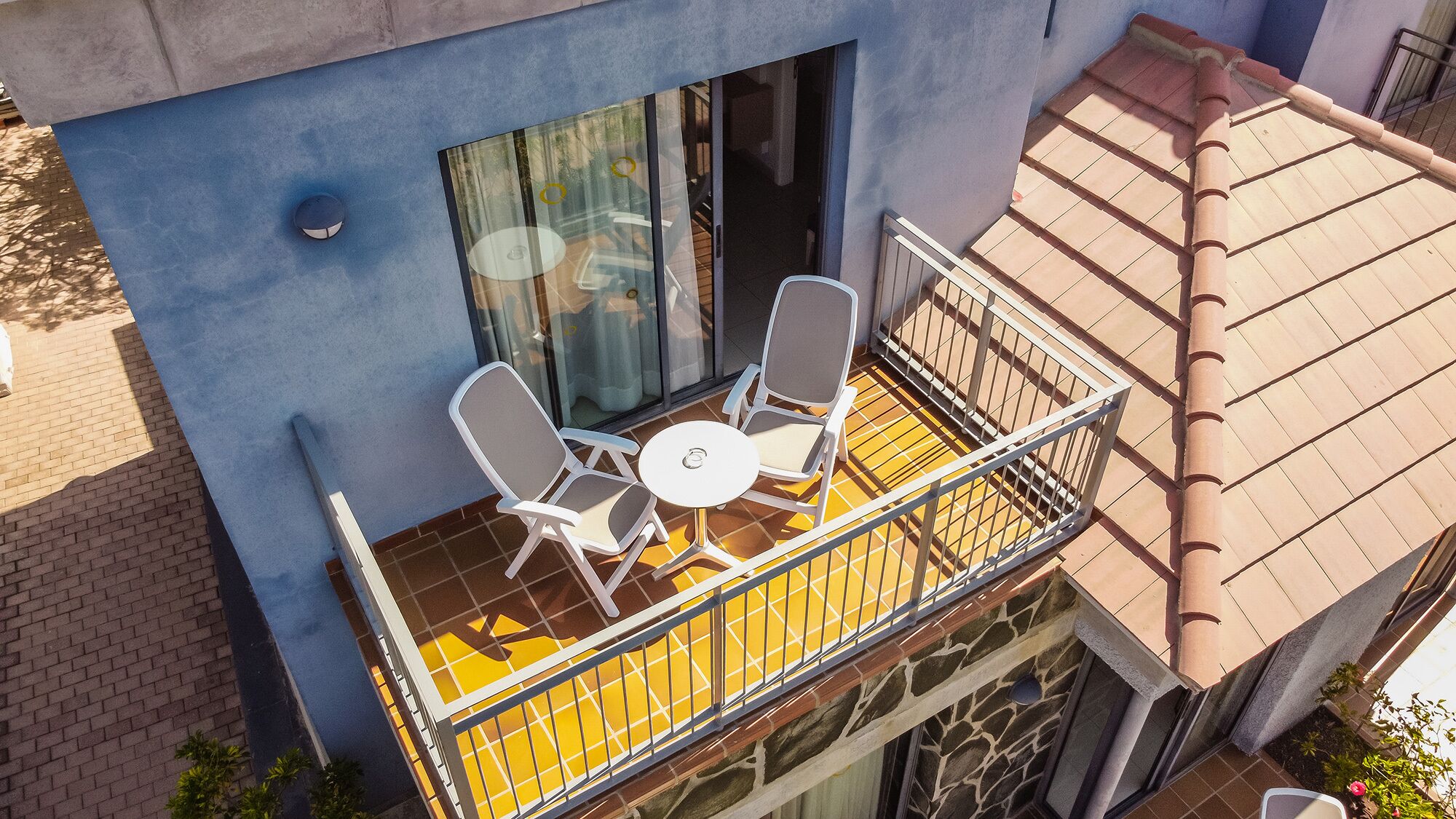 A balcony with two chairs and a small table, a window, and a tiled roof.