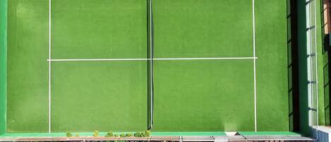 A tennis court with a net, surrounded by a green fence and a building with a brick facade.
