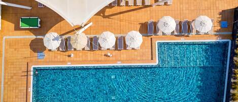 An aerial view of a swimming pool with adjacent sun loungers and umbrellas, a pool table, and a tiled deck area.
