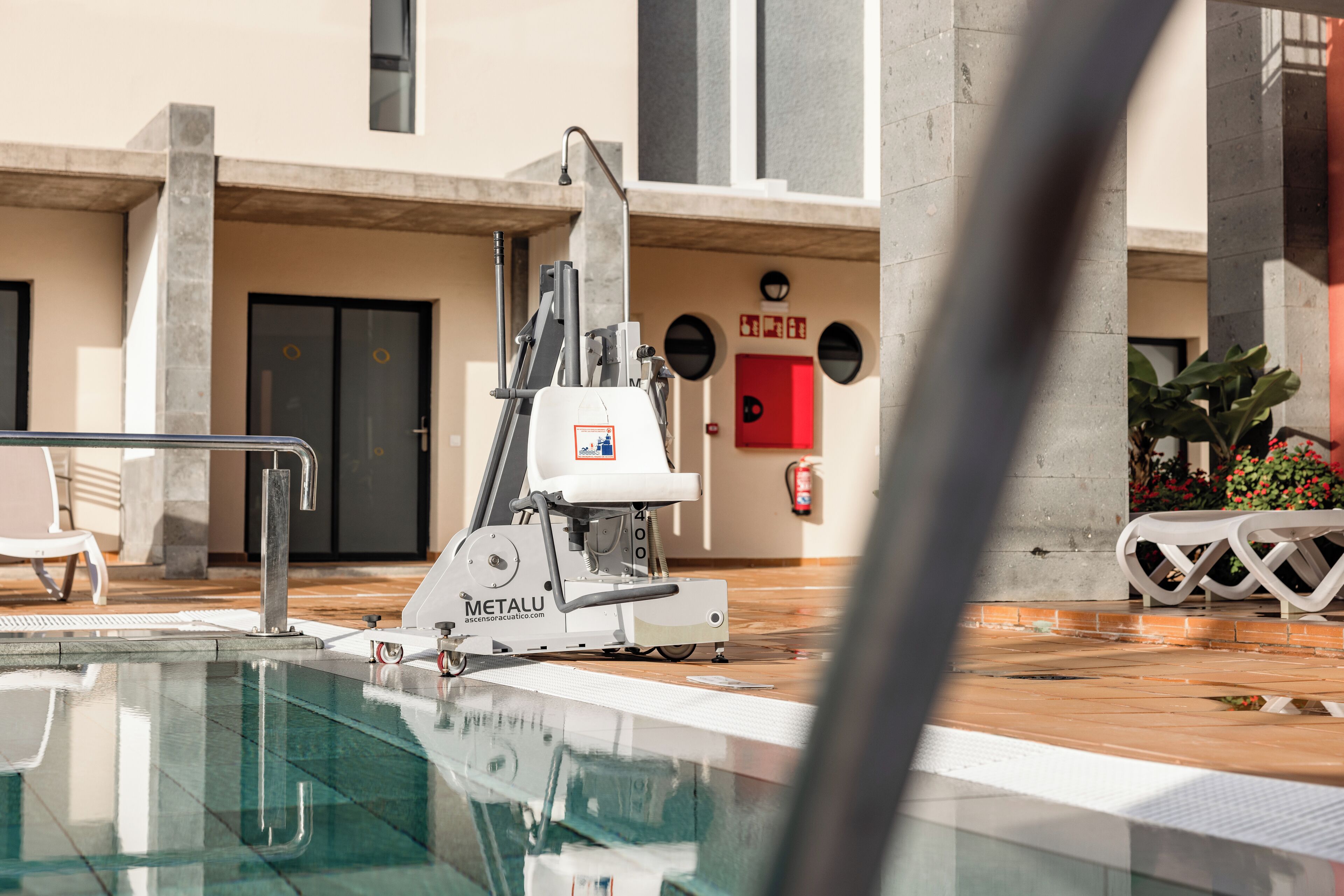 Zona de piscina con un limpiafondos automático, un extintor y una puerta de emergencia roja.