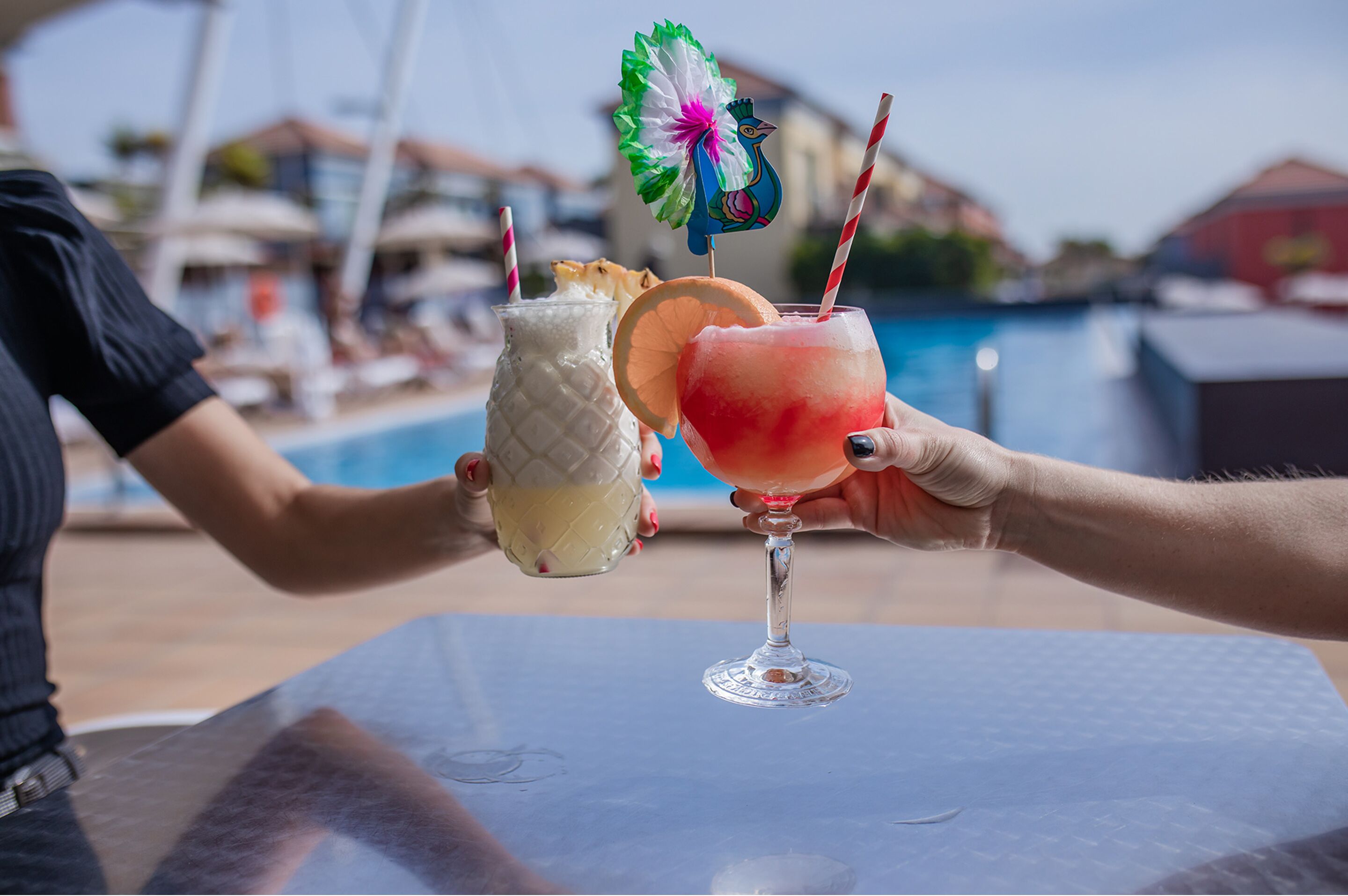 Dos personas sosteniendo bebidas en una mesa junto a la piscina, con una piscina y edificios al fondo.
