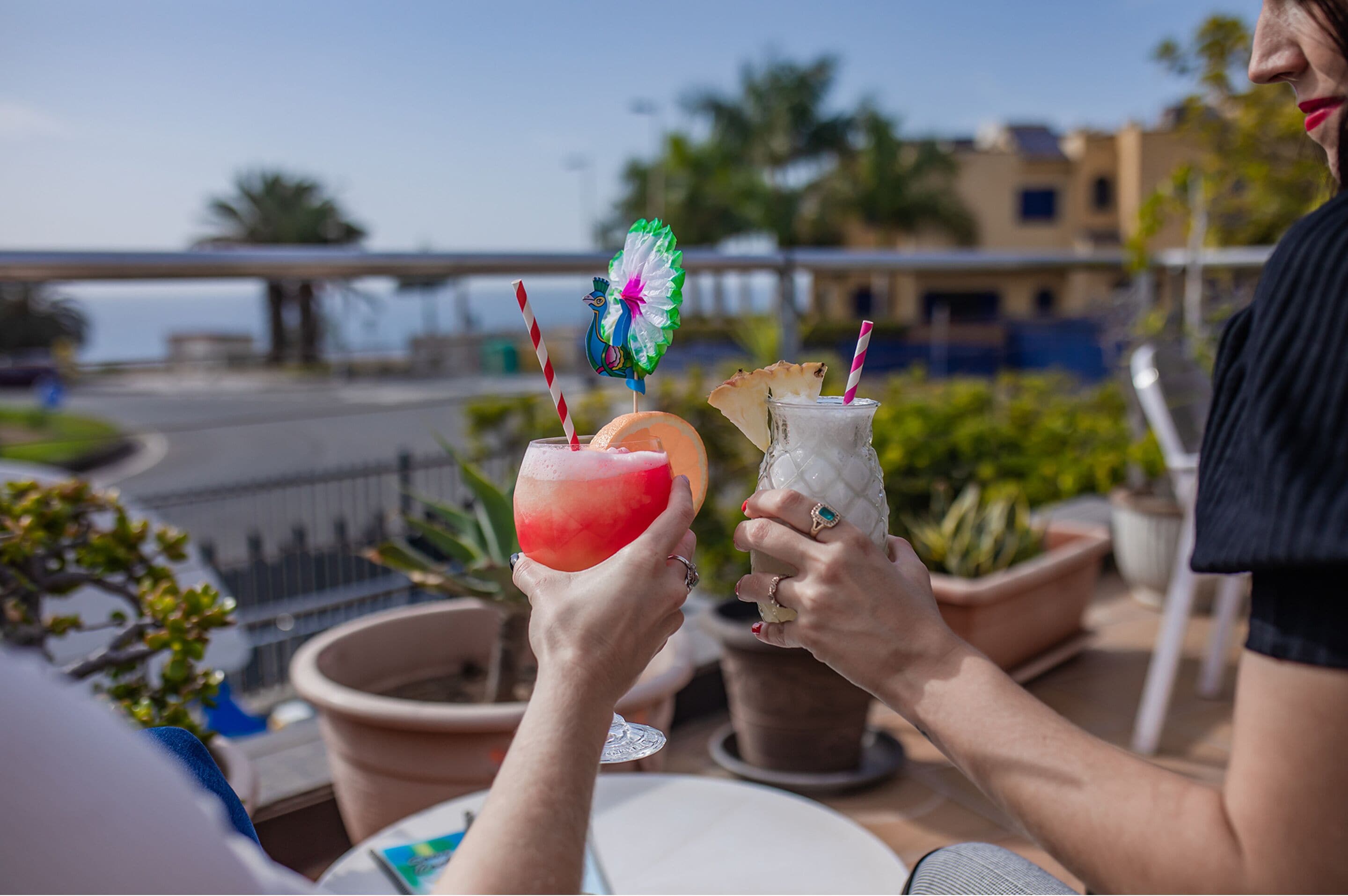 Two people holding drinks with straws and a decorative pinwheel, seated at an outdoor table with potted plants and a view of a bridge and buildings.