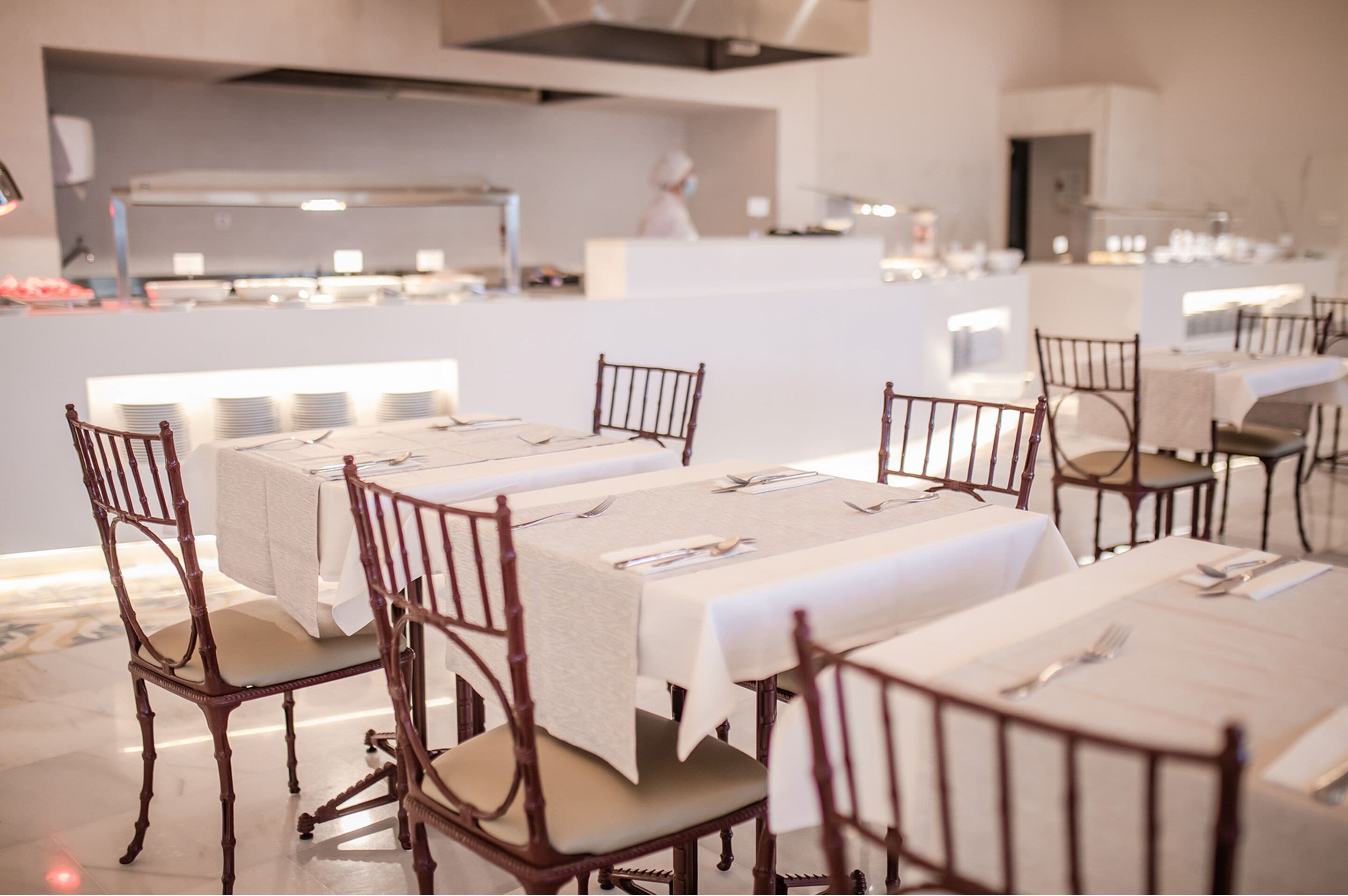 A dining area with tables set for service, chairs, and a kitchen in the background.