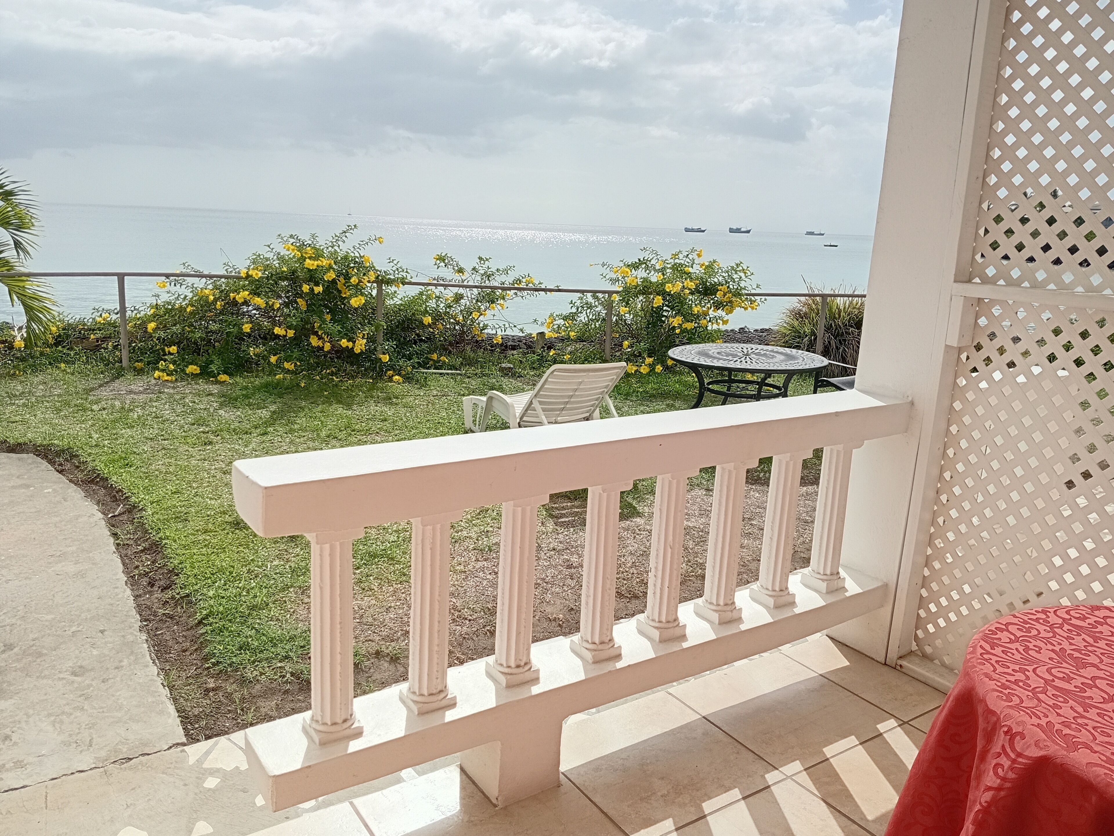 A balcony with a white railing, a small round table, and chairs, overlooking a body of water with boats and yellow flowering plants.