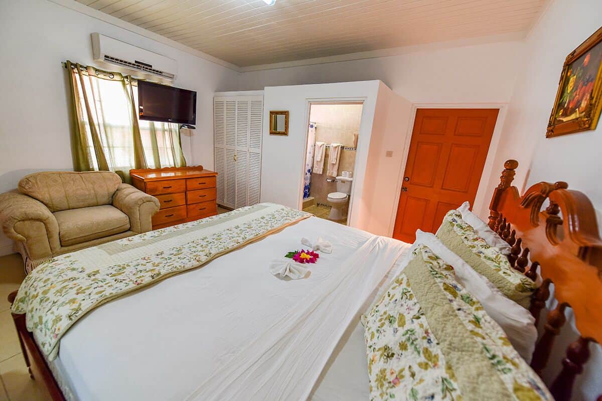 A bedroom with a bed, a chair, a dresser, a television, and a bathroom visible through an open door.