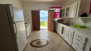 Una cocina con refrigerador, estufa, armarios y vista al océano a través de una puerta abierta.
