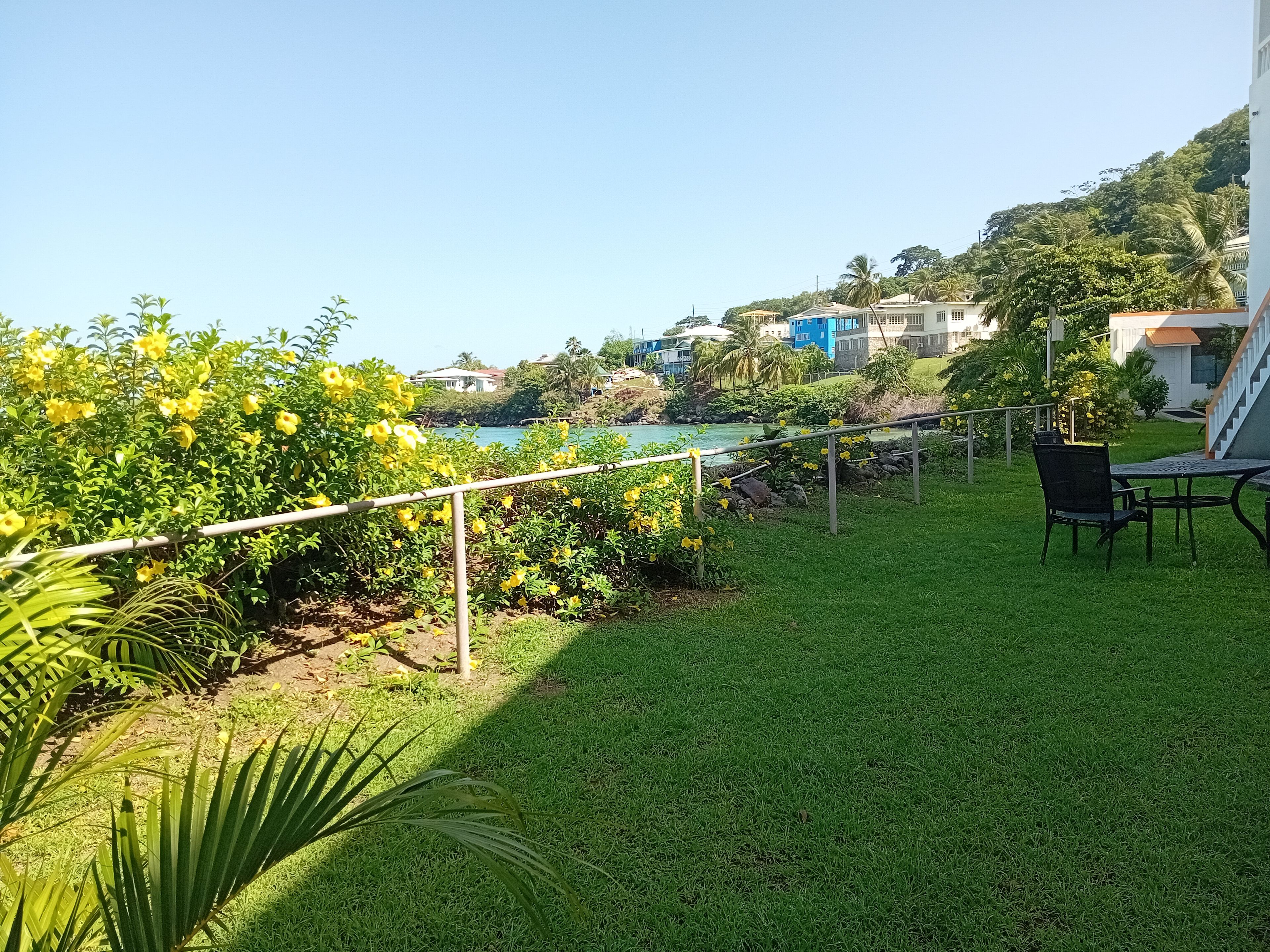 A grassy outdoor area with a table and chairs, a railing, and a view of a body of water and buildings in the distance.