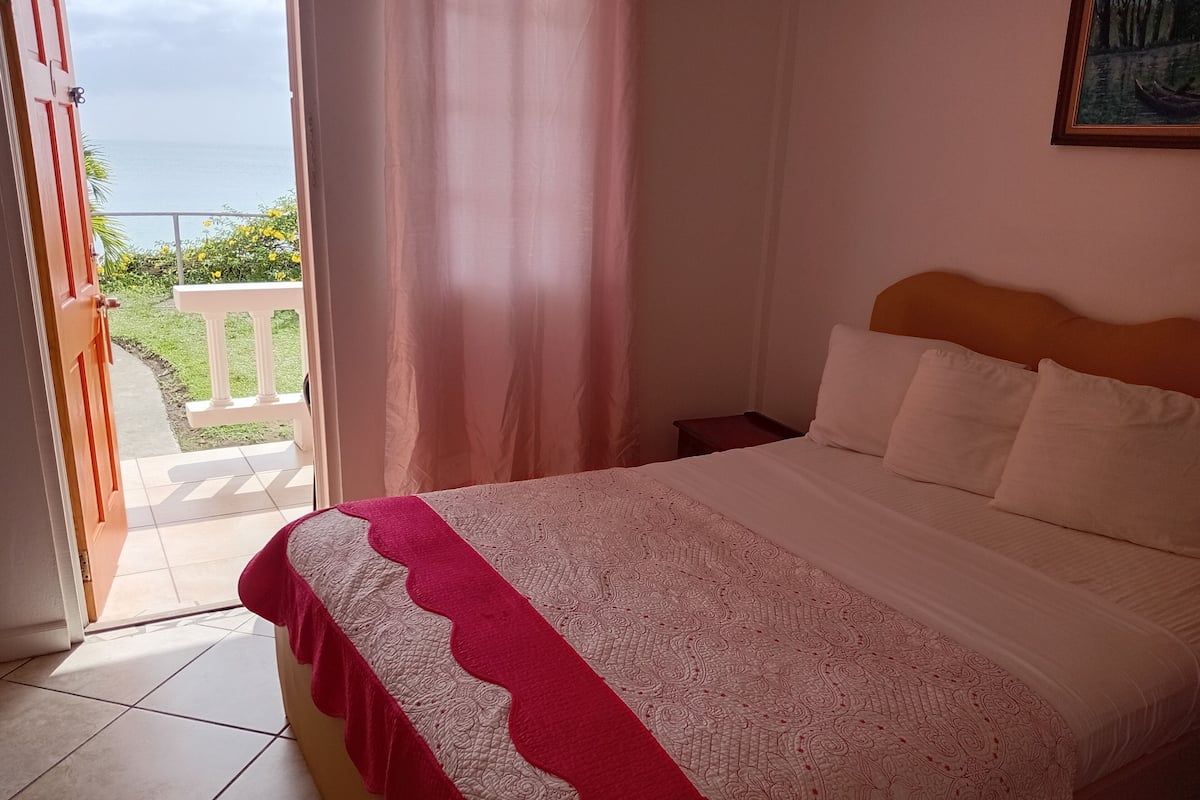 A bedroom with a bed, a nightstand, a window with curtains, and a view of the outdoors.
