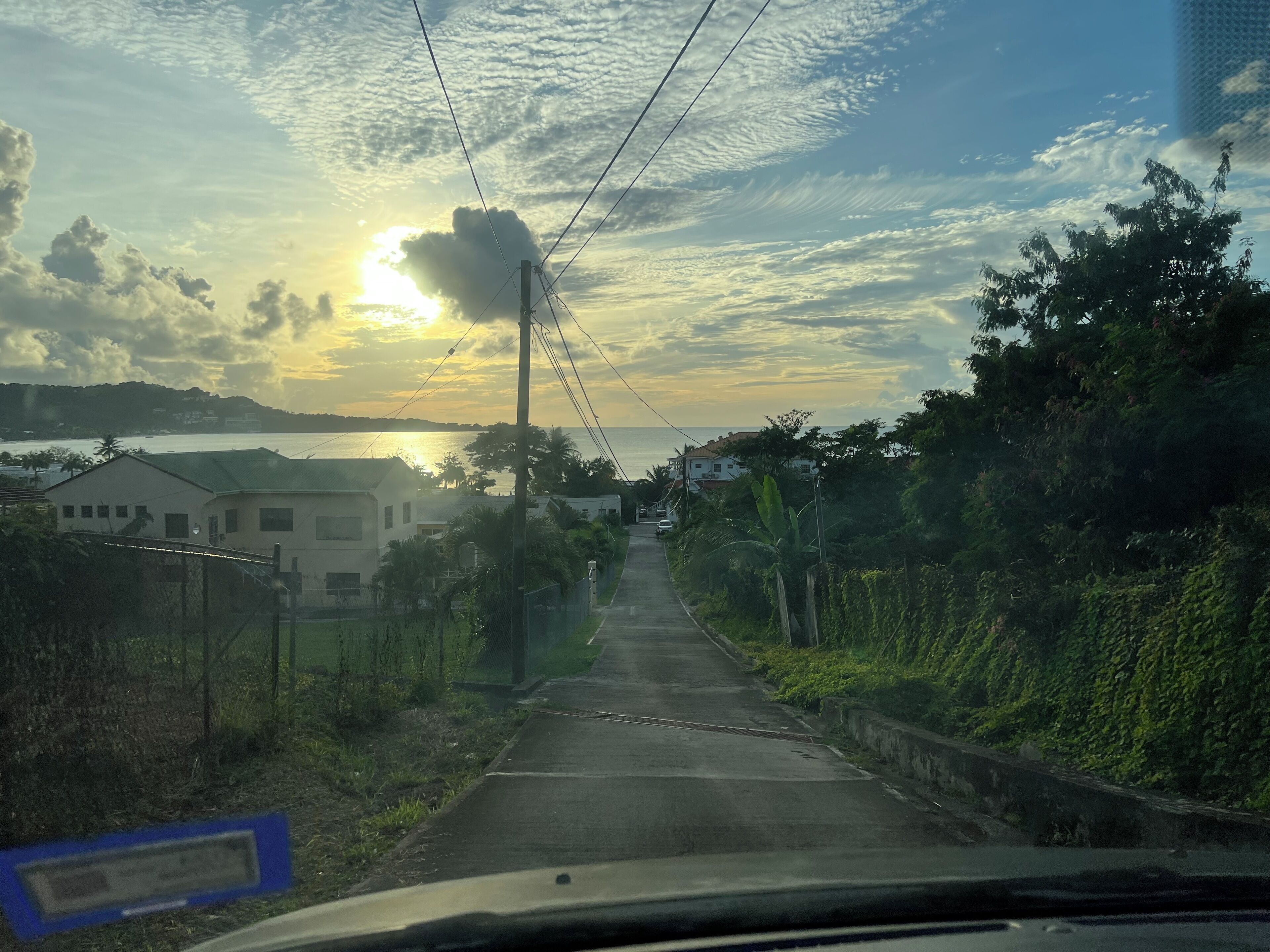 Una carretera que conduce a una zona residencial con casas a ambos lados, cielo despejado y el sol poniéndose al fondo.