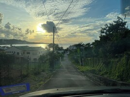 Una carretera que conduce a una zona residencial con casas a ambos lados, cielo despejado y el sol poniéndose al fondo.