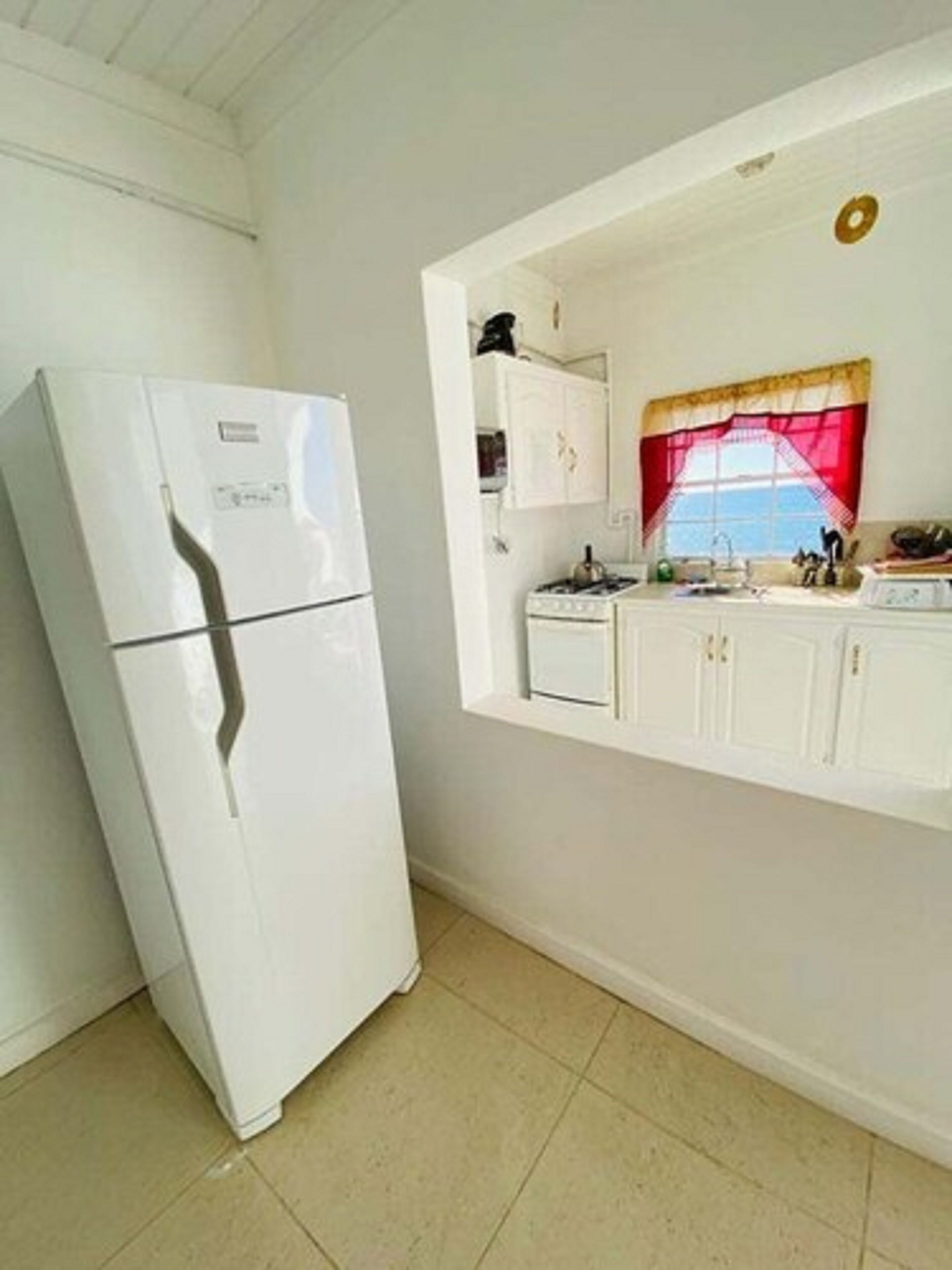 A compact kitchen with white cabinets, a white refrigerator, and a window with a red curtain.