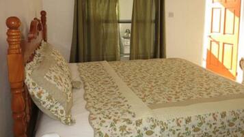 Un dormitorio con estructura de cama de madera, ropa de cama floral y vista a un baño a través de una ventana con cortinas verdes.