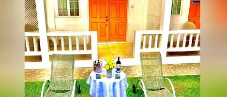 A covered outdoor seating area with two lounge chairs and a table, a double door entrance, and a small garden with grass and scattered leaves.