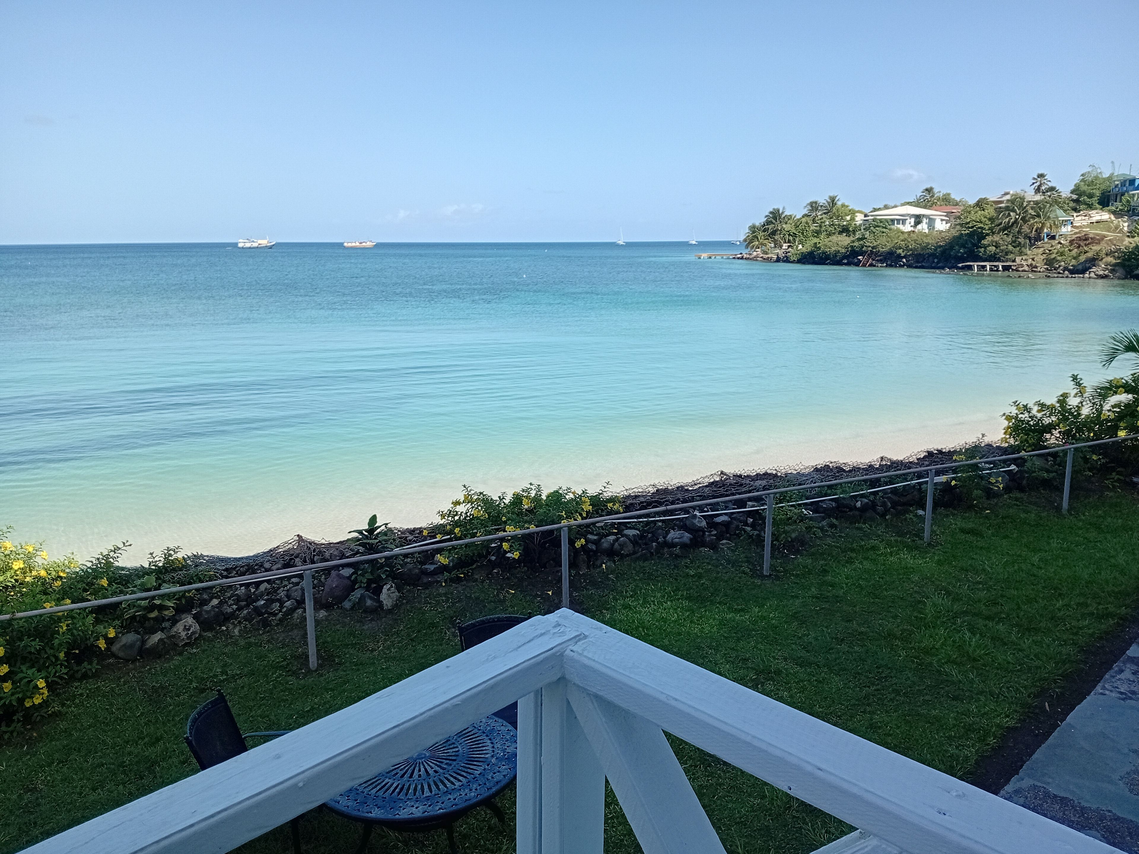 Vista de una playa con agua cristalina, un área con césped y una barandilla.