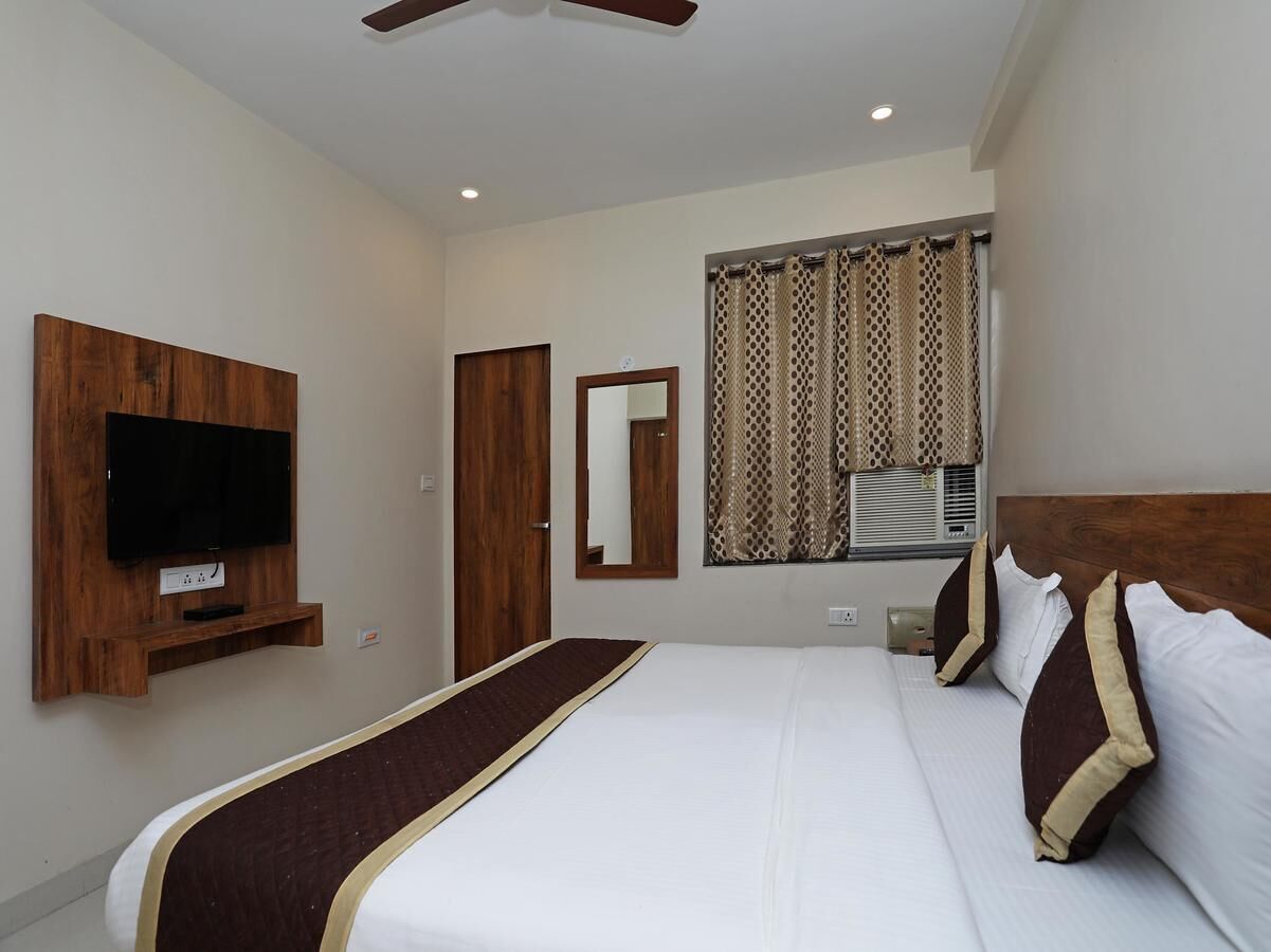 A hotel room with a large bed, a TV mounted on a wooden panel, a mirror, and a window with patterned curtains.
