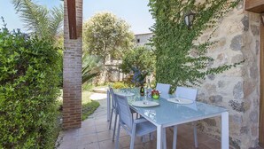 Un patio al aire libre con una mesa y sillas de vidrio, rodeado de vegetación y un muro de piedra.