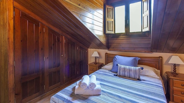 Un dormitorio con techo inclinado, paredes de madera, una cama grande con colcha a rayas, dos mesitas de noche con lámparas y una ventana con persianas.