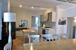 A modern kitchen with white cabinetry, a dining area with a wooden table and white chairs, and a window providing natural light. - The Apartment · Casa del Fattore, Villa Luzi (Marche)