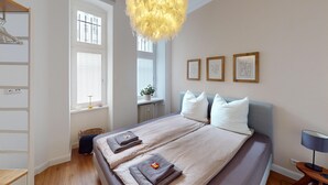 A bedroom with a bed, pillows, a bedside table, a lamp, a window, and a wall with framed pictures. - MyHappyplace in the middle of Berlin (Berlin)