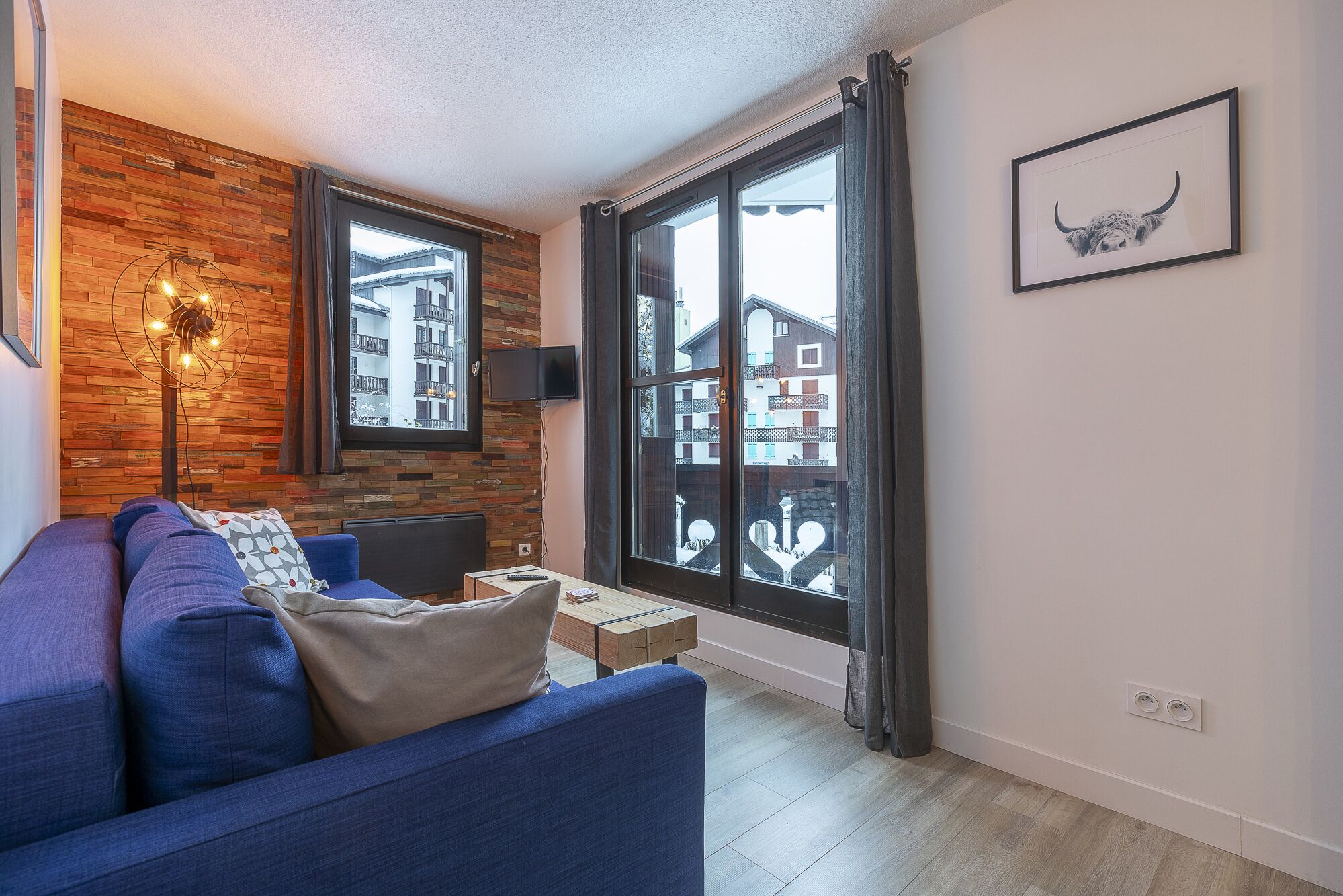 A modern living room with a blue sofa, a wooden accent wall, and a view of buildings through large windows.