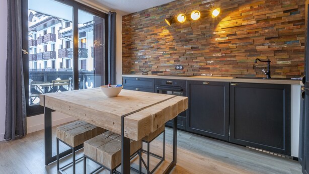 A modern kitchen with a wooden wall backsplash, a wooden dining table, and a view of the city through large windows. - Cham-Vue Apartment (Chamonix-Mont-Blanc)