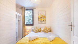A narrow room with a bed, yellow bedding, a window with a view of trees, and a painting on the wall. - Cham-Vue Apartment (Chamonix-Mont-Blanc)