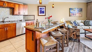Una cocina moderna con armarios de madera, salpicadero de azulejos y una isla central con taburetes de bar. Hay una zona de comedor con un sofá y un cuadro en la pared.