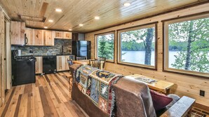 Interior de una cabaña con cocina, comedor y ventanas amplias con vista a un lago.