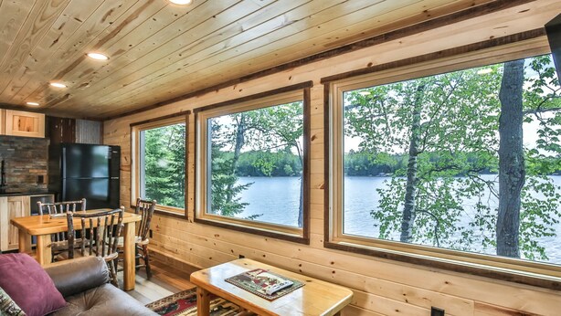 Una cabaña con comedor, sofá y grandes ventanales con vista a un lago.