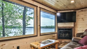 Interior acogedor de una cabaña con detalles de madera, una chimenea, un televisor y ventanas amplias con vistas a un lago.