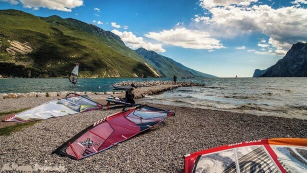 Een strand met vlieger en windsurfspullen, een rotsachtige kustlijn en bergen op de achtergrond.