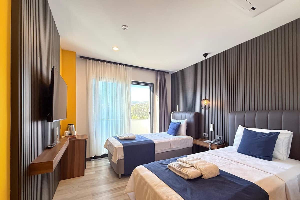 A modern hotel room with two beds, a flat-screen TV, a bedside table with a lamp, and a view of the outdoors.