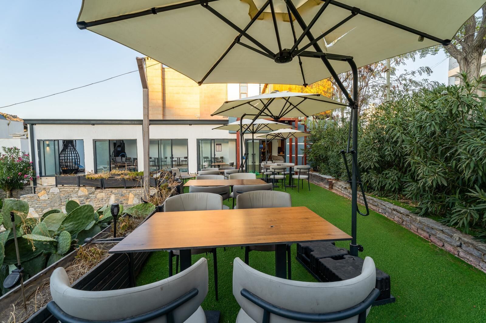 An outdoor seating area with tables and chairs under umbrellas, surrounded by greenery and a building in the background.