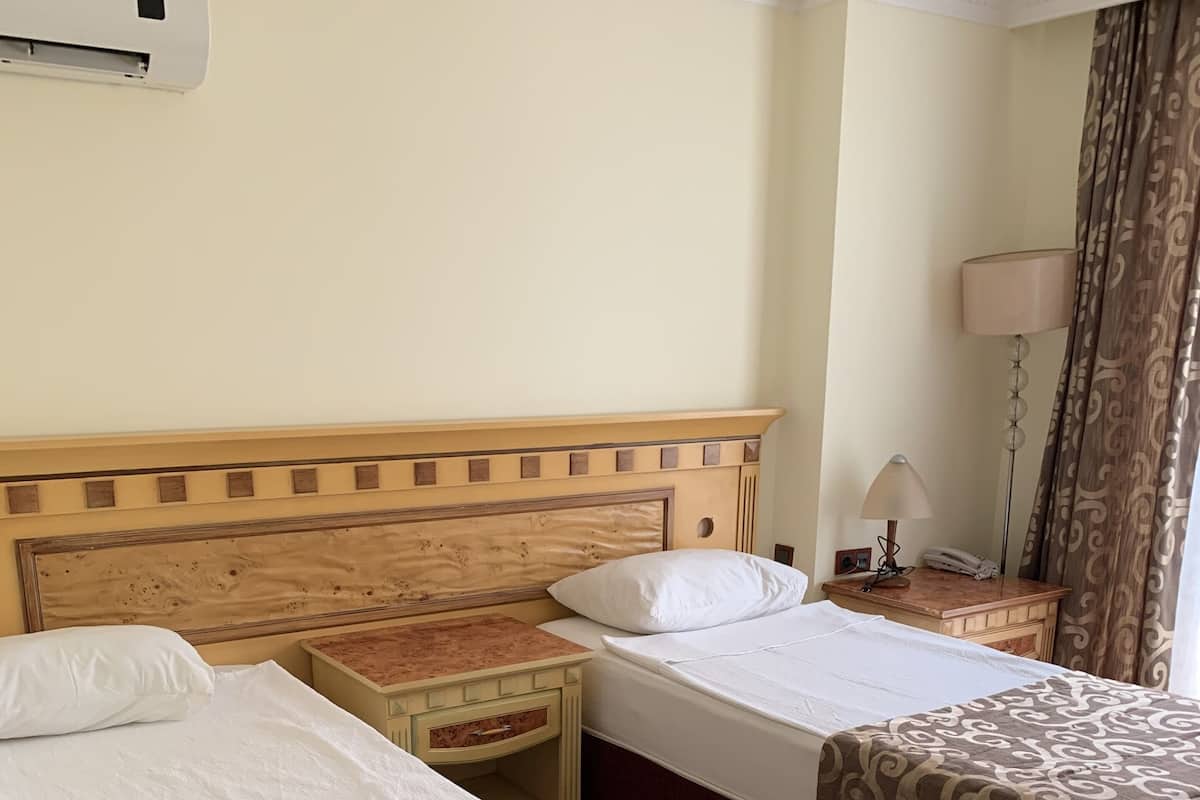 A hotel room with two beds, a bedside table with a lamp, an air conditioning unit, and a curtain.