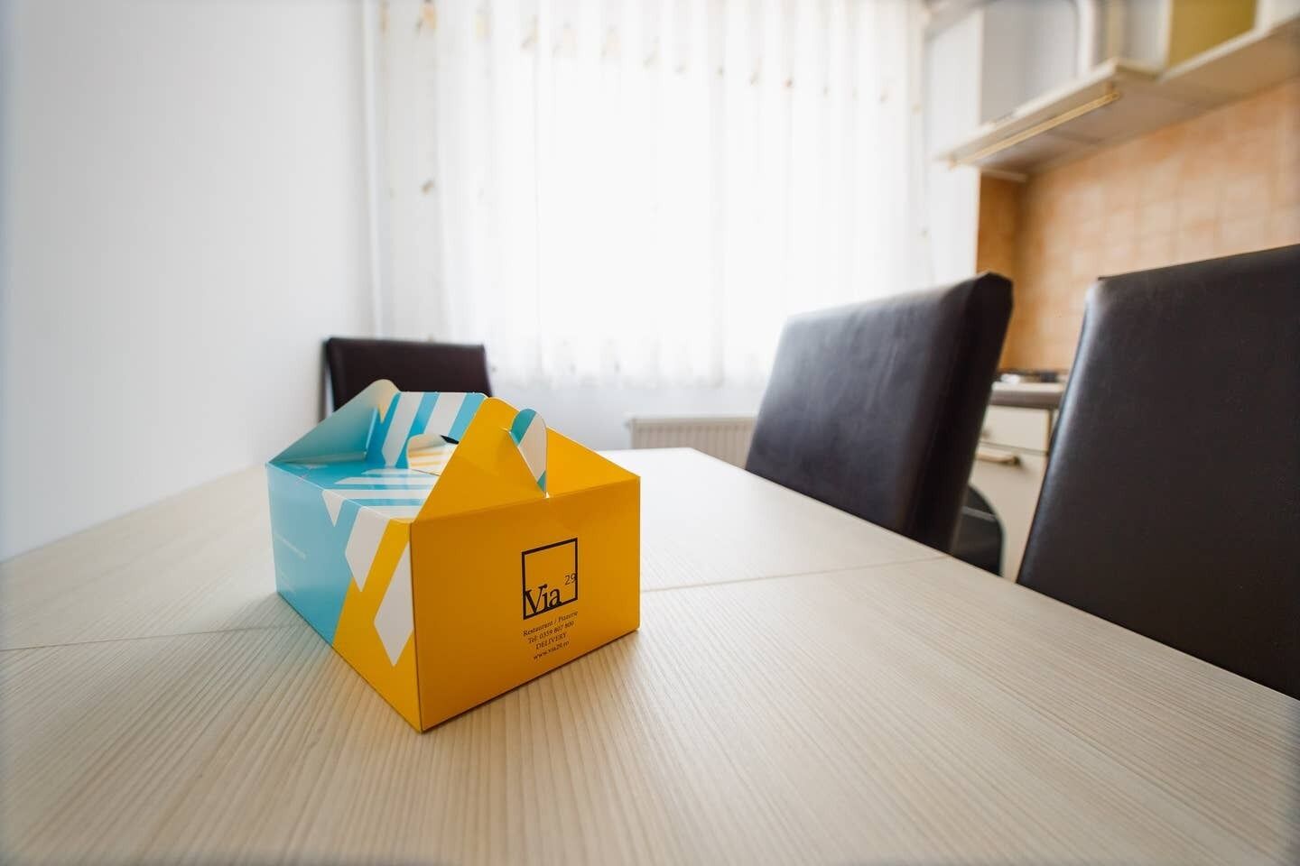 Een gele doos met een logo op een houten tafel in een kamer met een raam en stoelen.