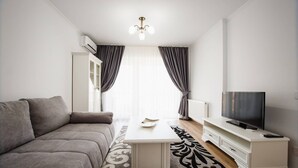 A modern living room with a grey sofa, a white coffee table, a flat-screen TV on a white stand, and a patterned rug. - Bright & Elegant Central Apartment (Oradea)