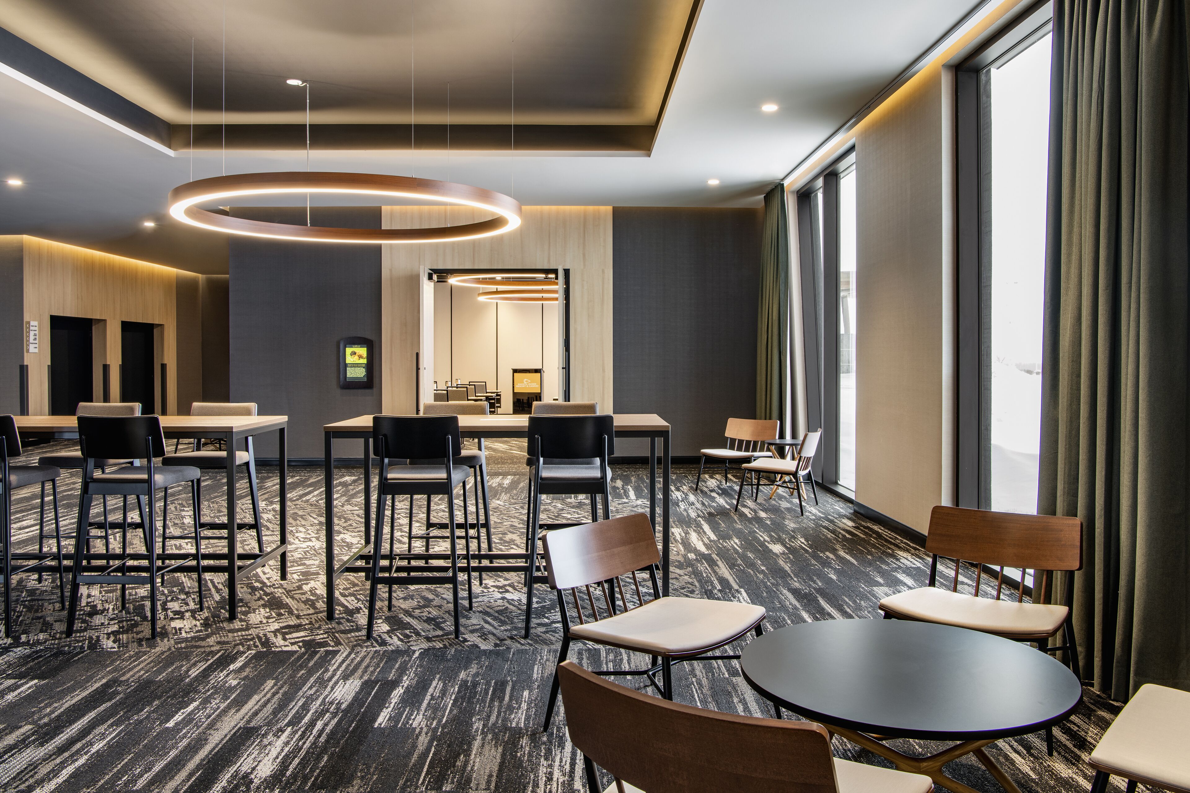 A modern hotel lobby with a carpeted floor, round tables, and chairs, a reception desk, and large windows with curtains.