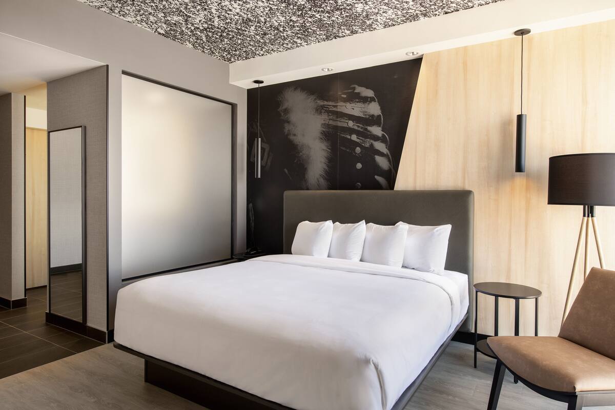 Una habitación de hotel moderna con una cama grande, una mesita pequeña, una silla y una obra de arte original en la pared.