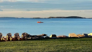 A coastal view with a row of wooden chairs facing a calm sea, a small boat in the distance, and a grassy area in the foreground. - Candy Cove 
Peace and seclusion by the Sea in Maine (Saint George)
