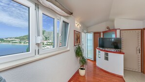 A room with large windows overlooking a scenic coastal view, a potted plant, a flat-screen TV, and a wooden floor. - Apartments Mia (Omis)