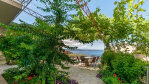 An outdoor patio with a table and chairs, surrounded by lush greenery and a view of the sea. - Apartments Mia (Omis)