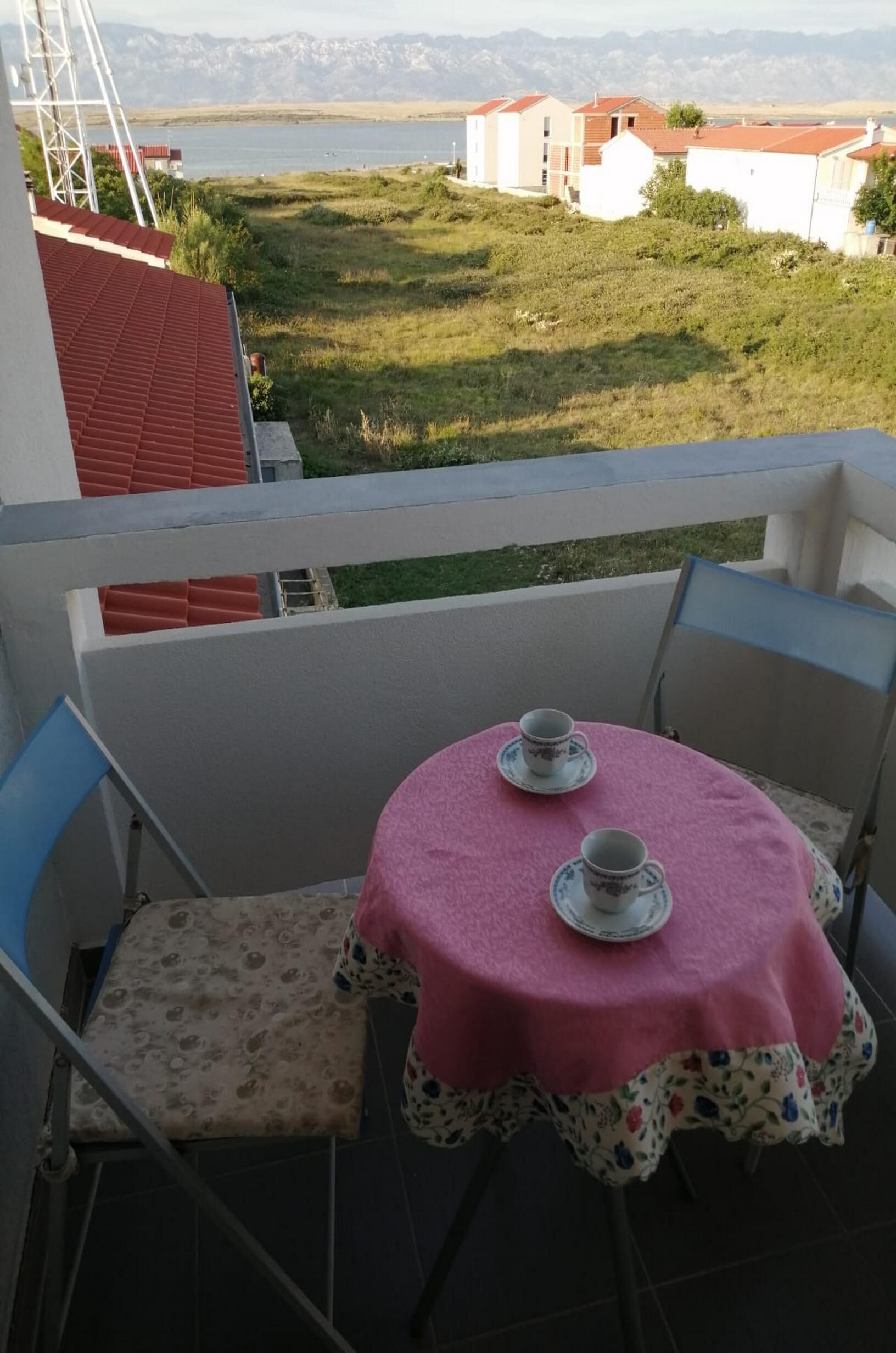 A balcony with a table set for two, overlooking a grassy area and a body of water with buildings in the distance.