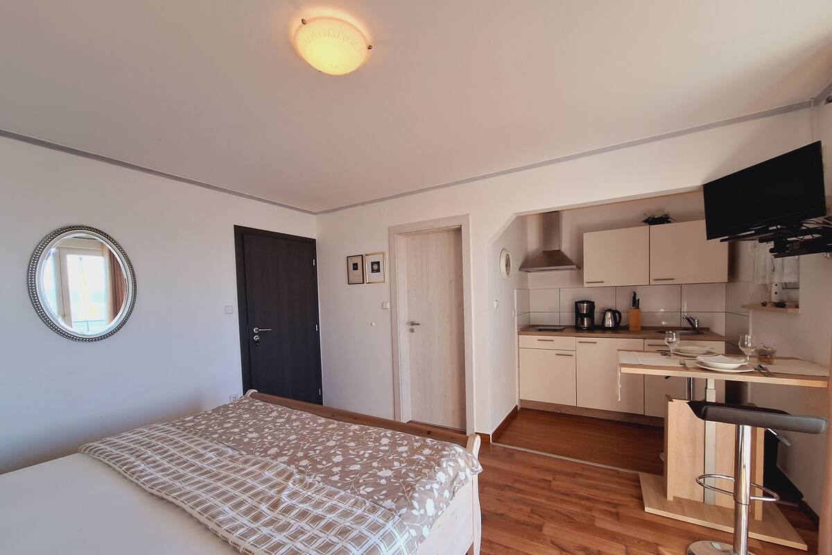 A compact room with a bed, a small kitchenette, a dining area, and a TV mounted on the wall.