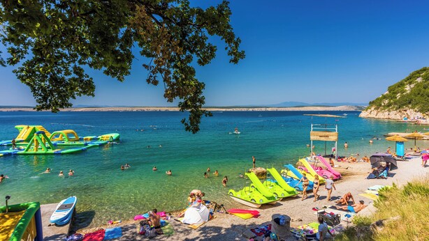 Ein Strand mit klarem Wasser, darin schwimmende Menschen und bunten Luftmatratzen.