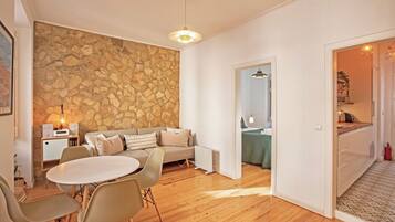 Uma sala de estar moderna com uma parede de pedra, um sofá, uma mesa de jantar com cadeiras, e uma cozinha visível através de uma porta aberta.