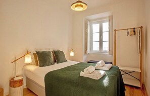 A hotel room with a bed, a window, a bedside table with a lamp, and a towel rack.