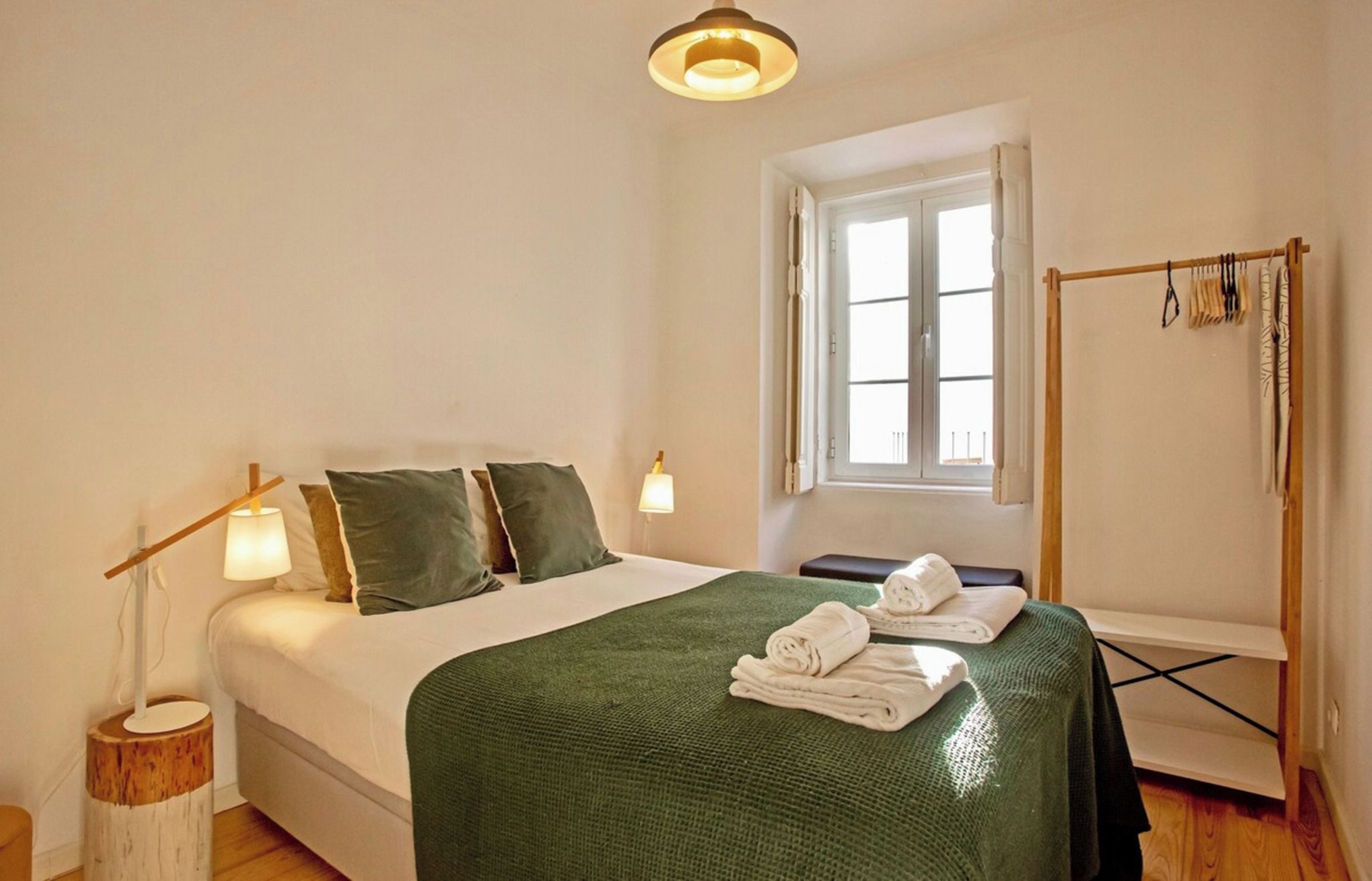 Una habitación de hotel con una cama, un ventanal, una mesita de noche con lámpara y un perchero para toallas.
